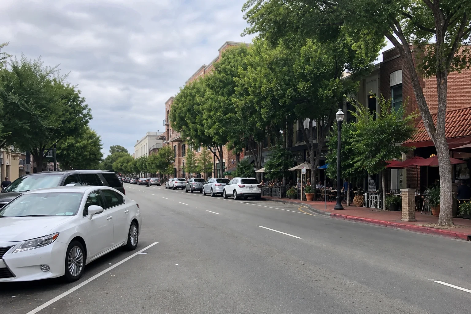 Tree-lined downtown main street with parked cars, brick storefronts, red awnings and outdoor café seating under an overcast sky