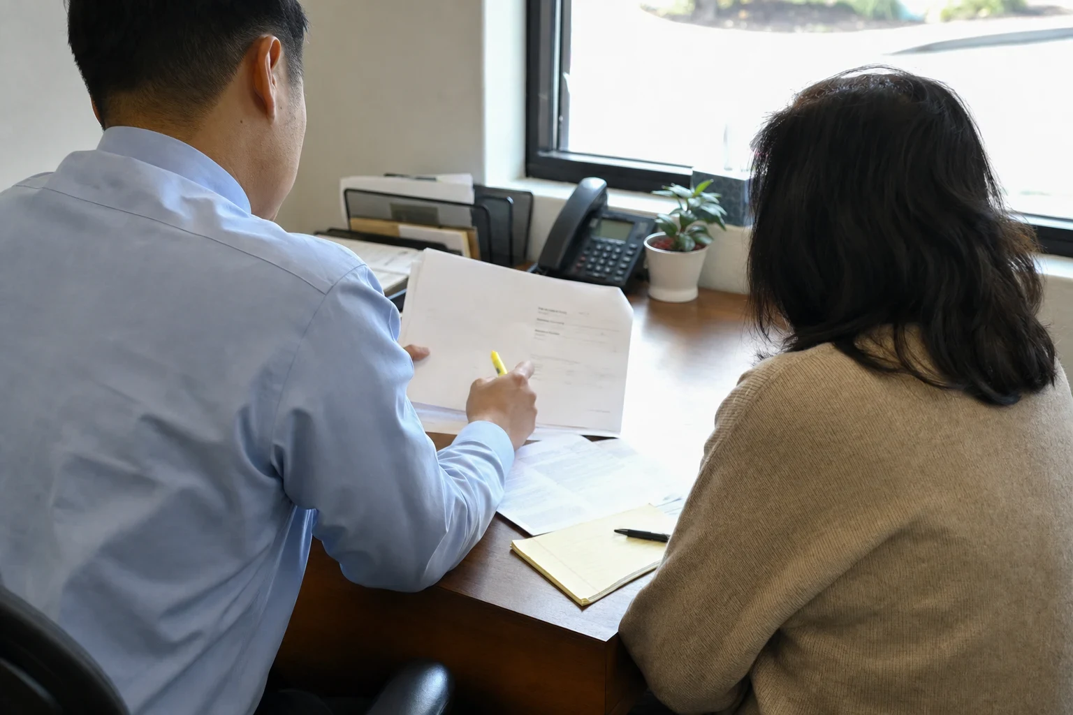 An agent in a light blue shirt reviews a document with a highlighter beside a woman in a tan sweater at an office desk with a phone, plant, notepad and window.